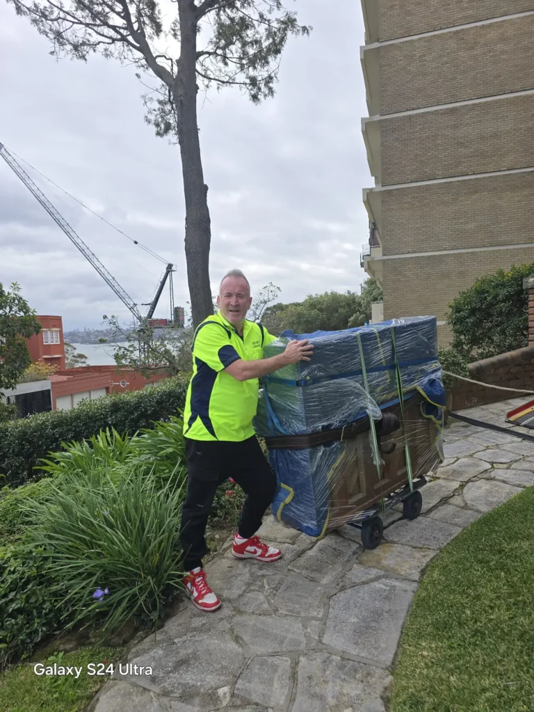 Anthony from Piano Pickup, a Sydney piano mover, with a wrapped upright piano strapped to a trolley on a stone path at a Point Piper property, Sydney Harbour in the background