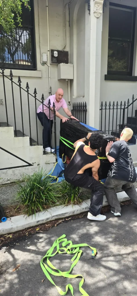 Three piano movers carefully lowering a shrink-wrapped upright piano down the front steps of a terrace home using lifting straps