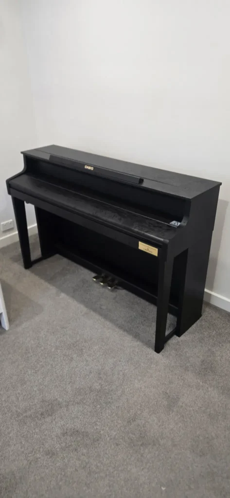 Black Casio digital piano with three pedals and slim cabinet stand, positioned against a wall on grey carpet