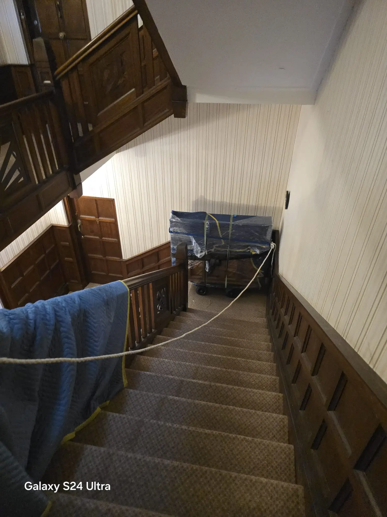 Upright piano wrapped in blue blankets and plastic on a piano trolley at the top of a carpeted staircase in a wood-panelled Point Piper home