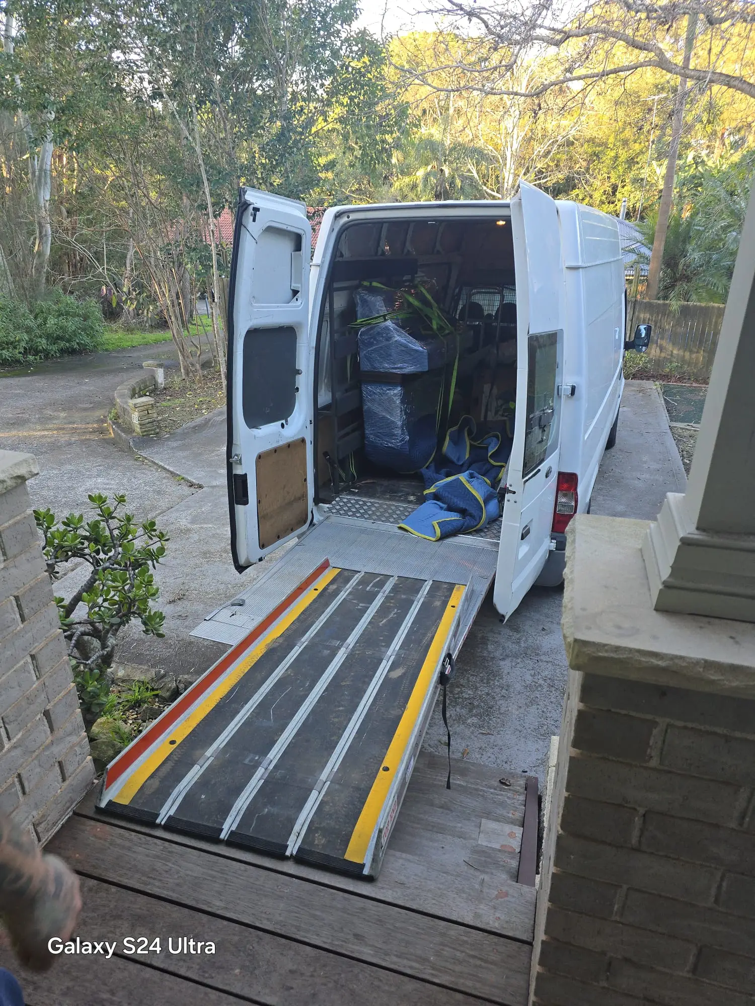 Piano Pickup van with rear doors open and the tail lift ramp lowered onto a Sydney driveway, wrapped piano and moving blankets visible inside