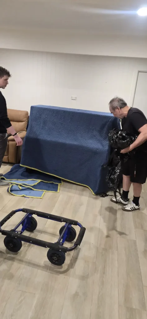 Two Piano Pickup movers covering an upright piano in padded moving blankets in a Sydney living room, with a four-wheel piano trolley waiting on the floor