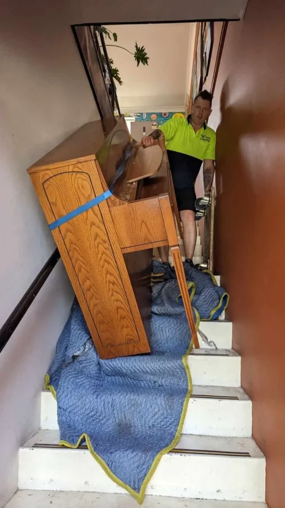 Piano Pickup mover in hi-vis guiding a strapped pianola down a narrow staircase with blankets protecting the treads and walls in a Pyrmont home