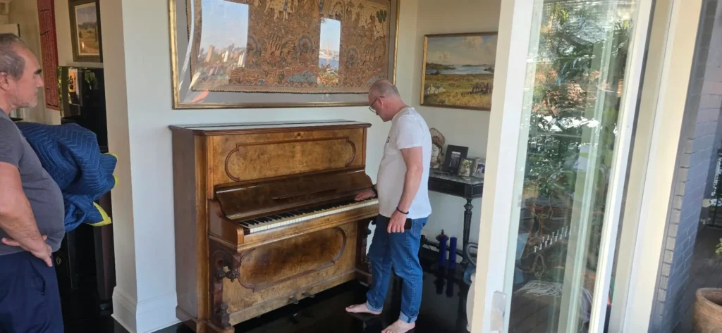 Older style walnut upright piano with carved detailing being inspected by two men in a living room before a piano move