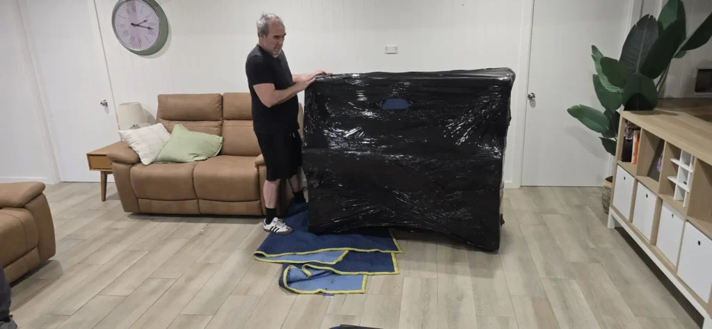 Piano Pickup mover sealing an upright piano in black protective plastic wrap over padded blankets, in a Sydney living room