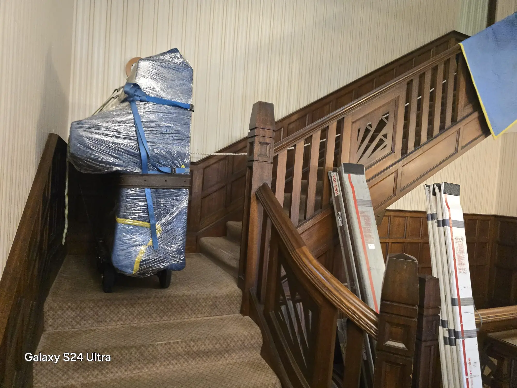 Piano fully wrapped in plastic and padded blankets, strapped to a piano trolley on a landing, with stair-climbing boards leaning against the timber banister